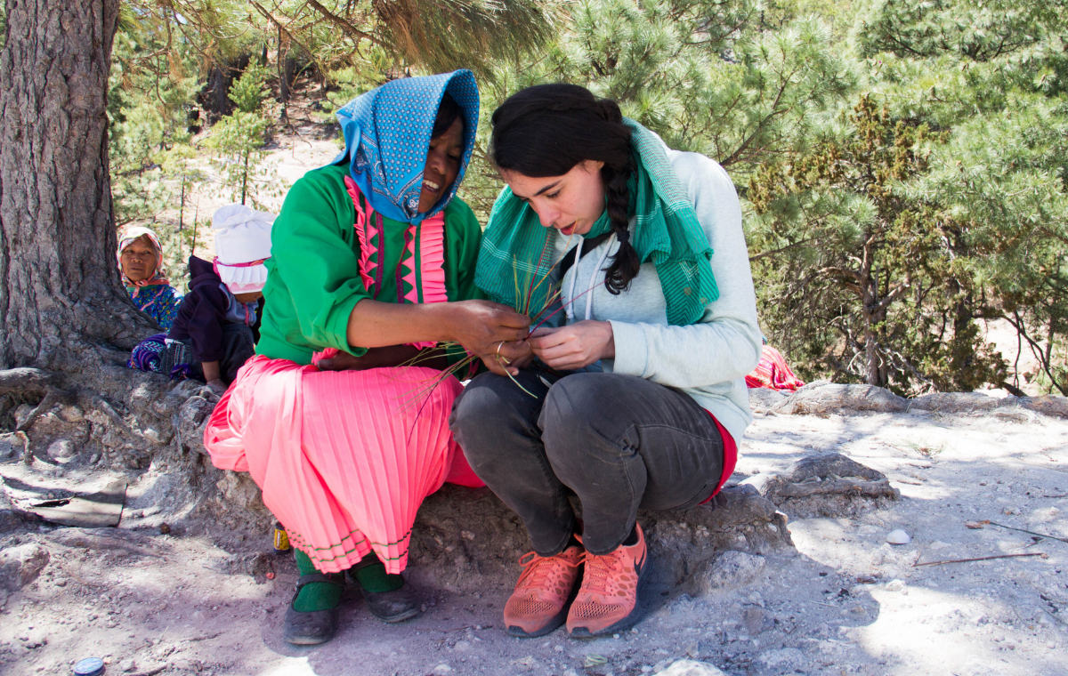 Two women sitting, Mexican woman in pink dress, green top, and blue headscarf helps tourist with weaving project.