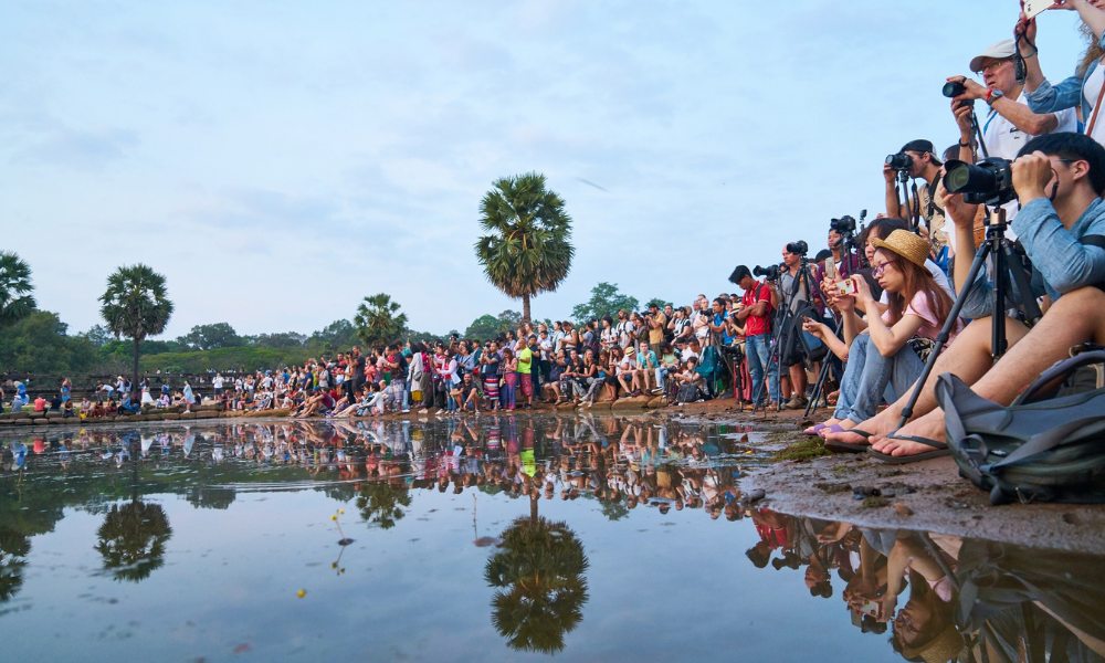 A horde of people in Cambodia gathered along the side of a large puddle, taking photos and looking towards a sight out of view of the photo.