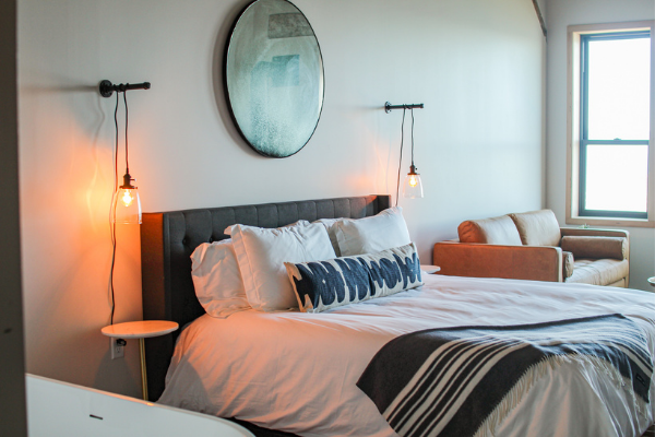 hotel bed with gray headboard, white sheets, and gray blanket draped over the edge of the bed. A caramel leather loveseat is in the corner of the room.