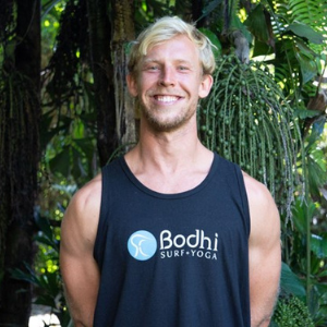 spencer-dunlap Blonde man smiling in front of tropical tree, wearing t-shirt with Bodhi Surf + Yoga logo
