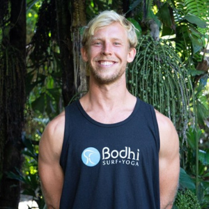 Blonde man smiling in front of tropical tree, wearing t-shirt with Bodhi Surf + Yoga logo