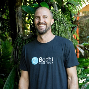 Man smiling in front of tropical tree, wearing t-shirt with Bodhi Surf + Yoga logo