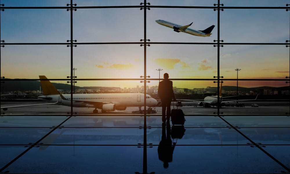 silhouette of man with luggage staring out airport window as plane flies into the air