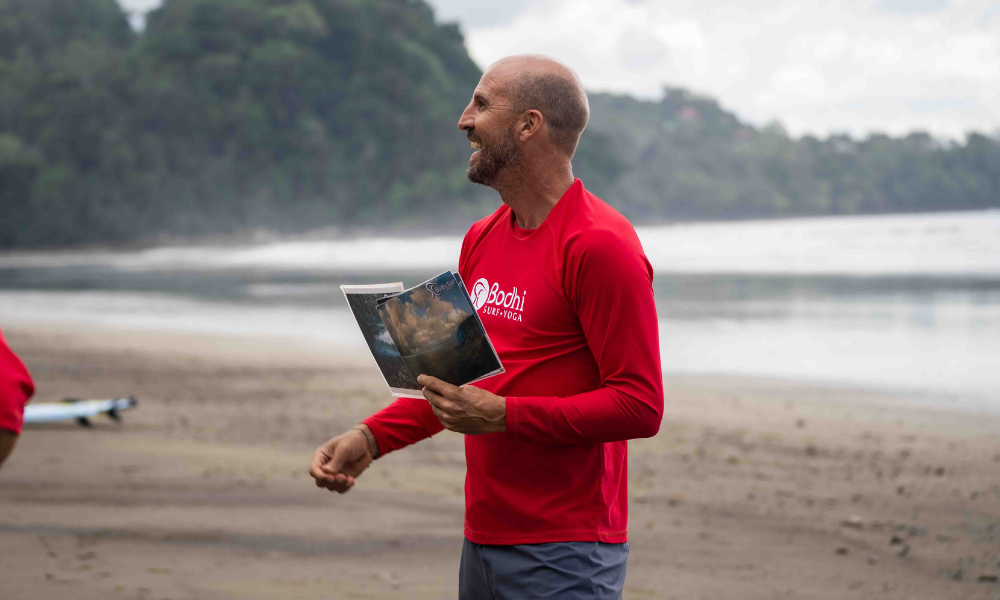 man in red long-sleeve shirt holding a piece of paper, smiling and looking to the left. He is standing on a tropical beach with trees behind him.