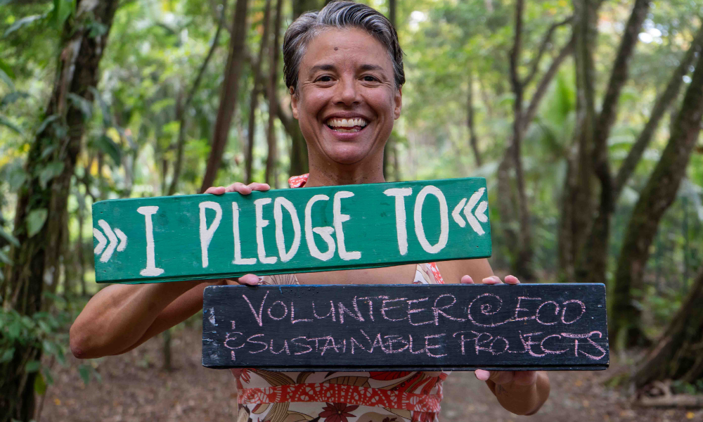 a smiling woman holding a sign that says "I pledge to volunteer at eco and sustainable projects."
