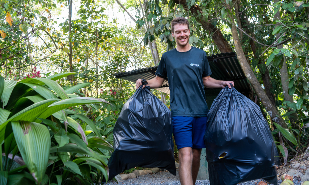 a man in a dark t-shirt holding two black trash bags in the jungle.
